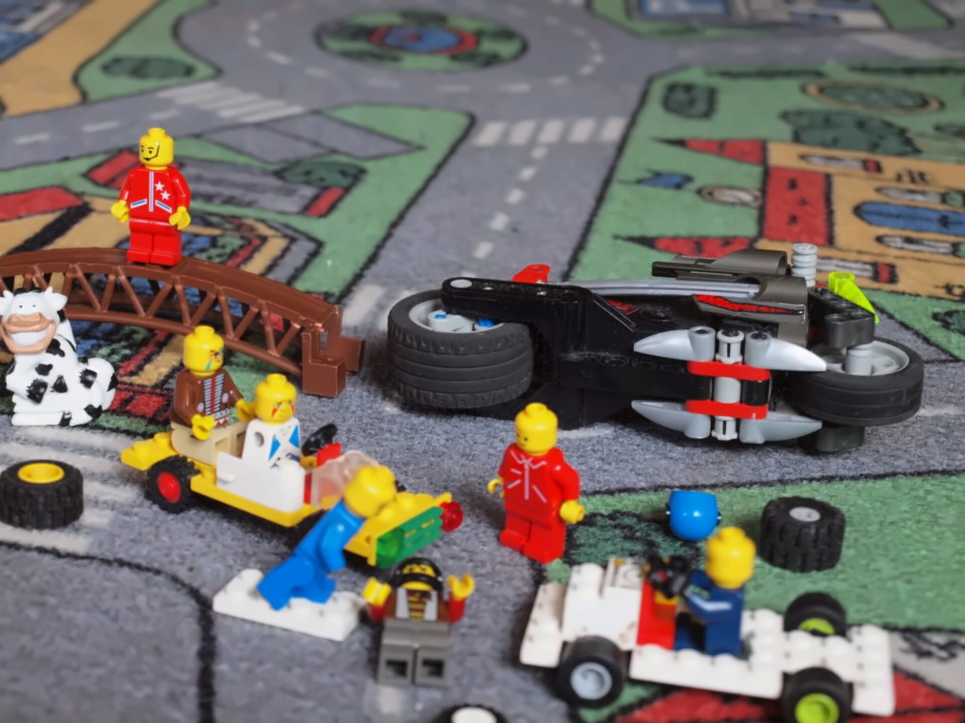 Nachgestellter Verkehrsunfall mit Legofiguren auf einem Spielteppich: ein umgekipptes Motorrad, ein Zuschauer auf einer Brücke, eine Kuh am Straßenrand, verstreute Räder und ratlose Beteiligte